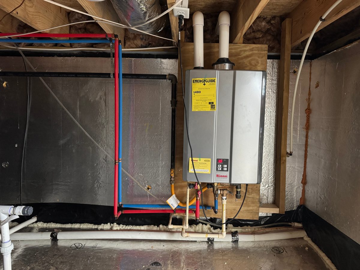 A propane tankless water heater mounted on a plywood panel in a sealed crawlspace, with red and blue water lines and a black gas pipe beside it
