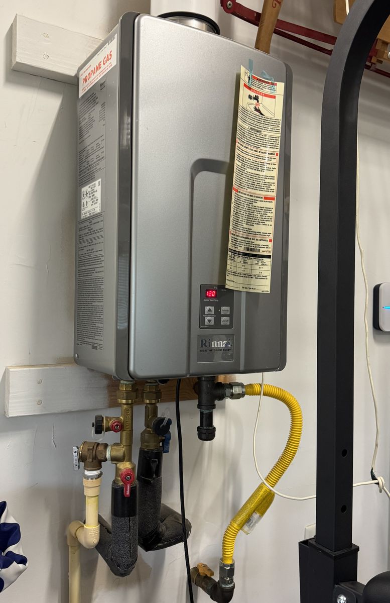 A propane tankless water heater on a garage wall with its gas connection and water lines below