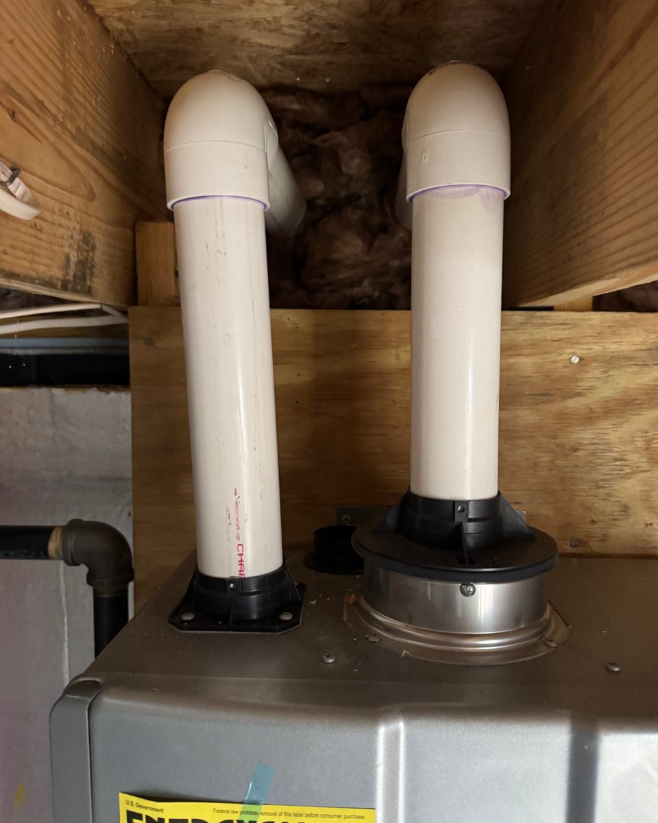 Two white plastic vent pipes rising from the top of a tankless unit and turning into the floor framing of a crawlspace