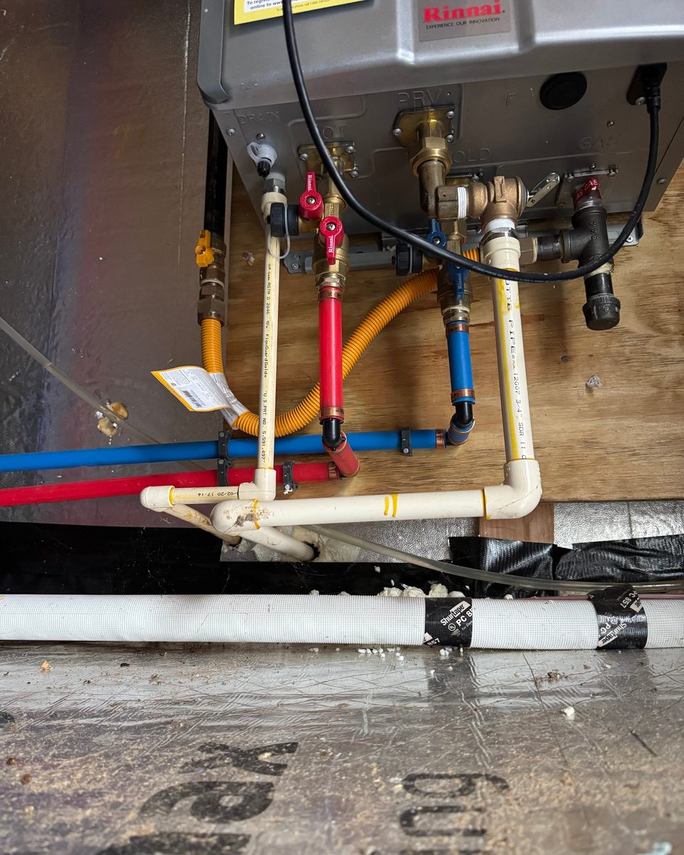 View from below of the service valves, pressure relief valve, drain lines, and gas connection on a crawlspace tankless unit